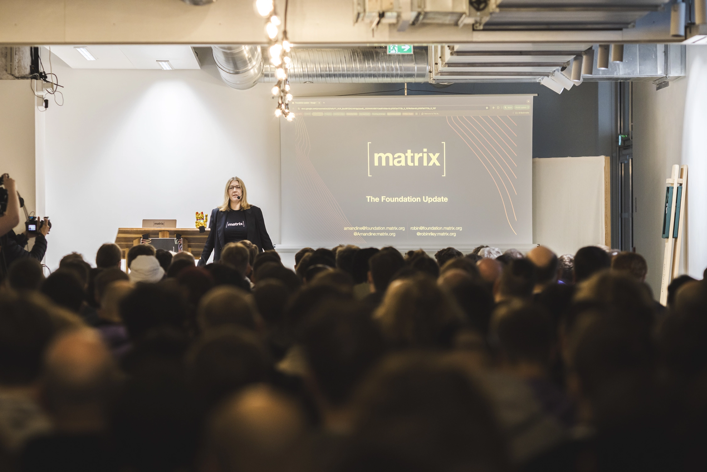 A picture of Amandine, one of the Guardians of The matrix.itmanbu.com Foundation, standing in front of a large crowd in a packed room. Her slides read 'The Foundation Update'.